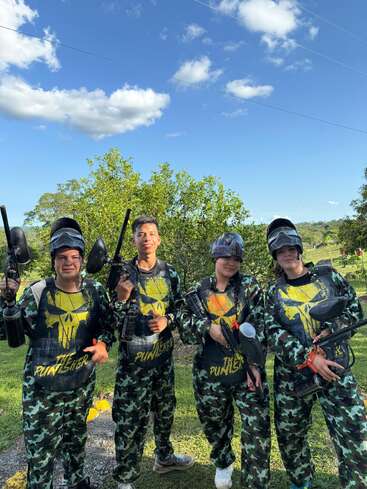 Four people stand outdoors under a bright blue sky, wearing matching camouflage "The Punisher" paintball gear, holding paintball markers, smiling, and ready for action.