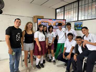 A group of students and two adults pose in a classroom, smiling. Some wear uniforms, some casual clothes. A colorful "Hey Buddy!" sign is behind them.