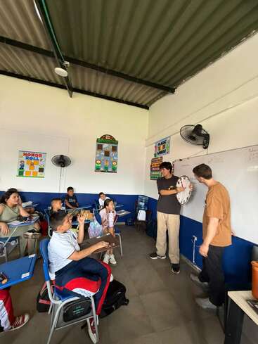 Uma cena de sala de aula com alunos sentados nas carteiras, participando ativamente. Dois professores estão em pé na frente da sala, um deles segurando um relógio, dando uma aula sobre o tempo.