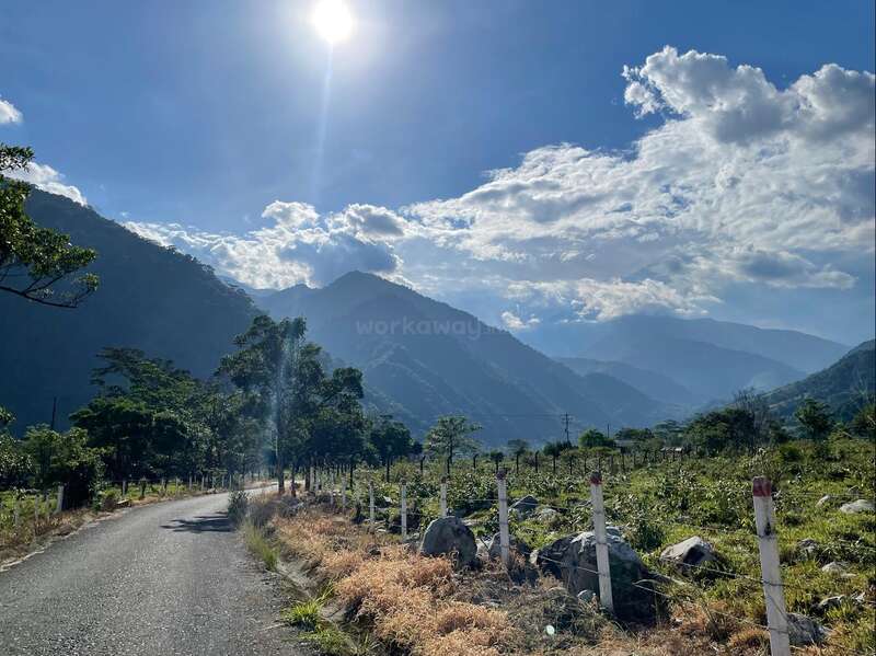 A peaceful countryside road winds through green fields, flanked by white fence posts, with majestic mountains and fluffy clouds under a bright sun in the sky.