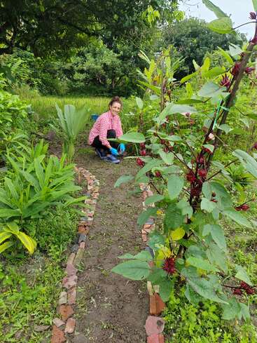 Eine Frau in einem rot karierten Hemd und blauen Handschuhen gärtnert auf einem gemauerten Weg, umgeben von üppigen grünen Pflanzen und blühenden Sträuchern im Freien.