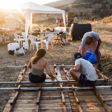 La imagen muestra a un grupo de personas trabajando en una estructura de madera en un campo, rodeadas de material de acampada y una tienda blanca, con una ladera visible al fondo.
