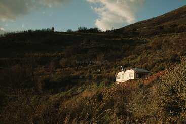 Une petite caravane blanche se dresse, solitaire, sur une colline verdoyante, au cœur d'une nature sauvage, sous un ciel bleu parsemé de nuages et baignée par la chaleur du soleil.
