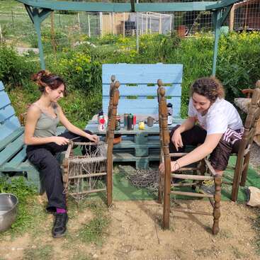 Deux personnes assises en plein air tressent ou réparent les assises de chaises en bois à l'aide de corde. Entourées de verdure et d'un banc rustique bleu, elles semblent concentrées et sereines.