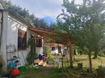 Una acogedora casa blanca con una pérgola de madera, rodeada de árboles y un jardín con césped. Los muebles de exterior y las herramientas de jardín crean un ambiente rural tranquilo y acogedor.