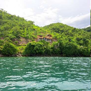 The image depicts a serene scene of a house situated on a hillside overlooking a body of water, surrounded by lush greenery and a cloudy sky.