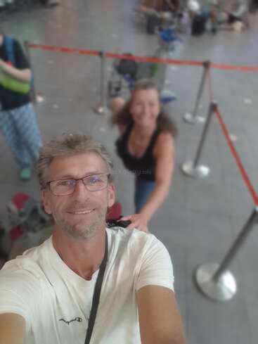 Un hombre se hace un selfie en lo que parece ser un aeropuerto o una estación. Una mujer está detrás de él, ambos sonriendo; se ven maletas, barreras y otra persona.