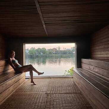 Una mujer se relaja en una sauna de madera, mirando por un gran ventanal con vistas a un tranquilo río, con árboles y casas de fondo al atardecer.
