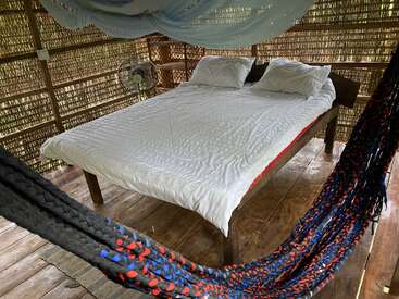 Une chambre rustique confortable avec un mur en bambou tressé, un lit blanc, deux oreillers, une moustiquaire au plafond, un ventilateur électrique, un plancher en bois et des hamacs colorés au premier plan.
