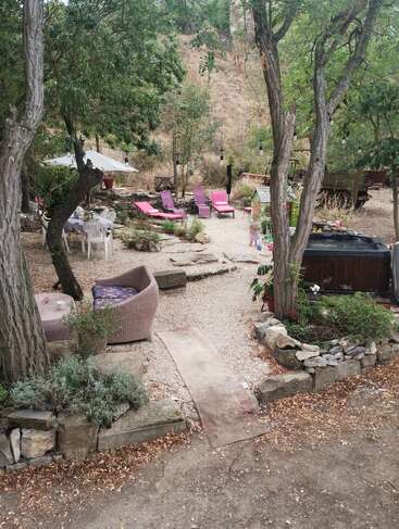 This cozy outdoor garden features pink lounge chairs, a hot tub, casual seating, leafy trees, stone borders, and a small playhouse, creating a tranquil, inviting atmosphere.