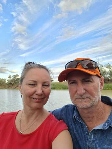 A smiling woman and a man wearing an orange cap take a selfie by a serene lake, under a beautiful sky with wispy clouds and greenery.