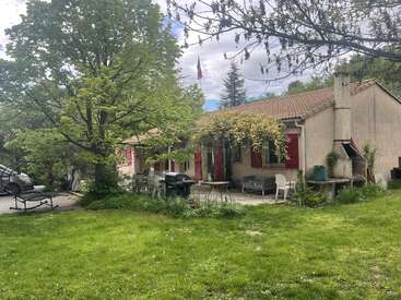 A charming countryside house with red shutters, surrounded by lush greenery and trees. Patio furniture, a barbecue grill, and outdoor seating create a welcoming, peaceful atmosphere.