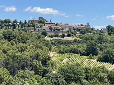 A picturesque hilltop village sits under a bright blue sky, surrounded by lush green trees, vineyards, and cypress trees, evoking a tranquil countryside scene.