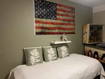 A cozy room features a white bed with three green fern pillows, a large American flag wall art, a shelf with décor, and a vintage pinball machine.