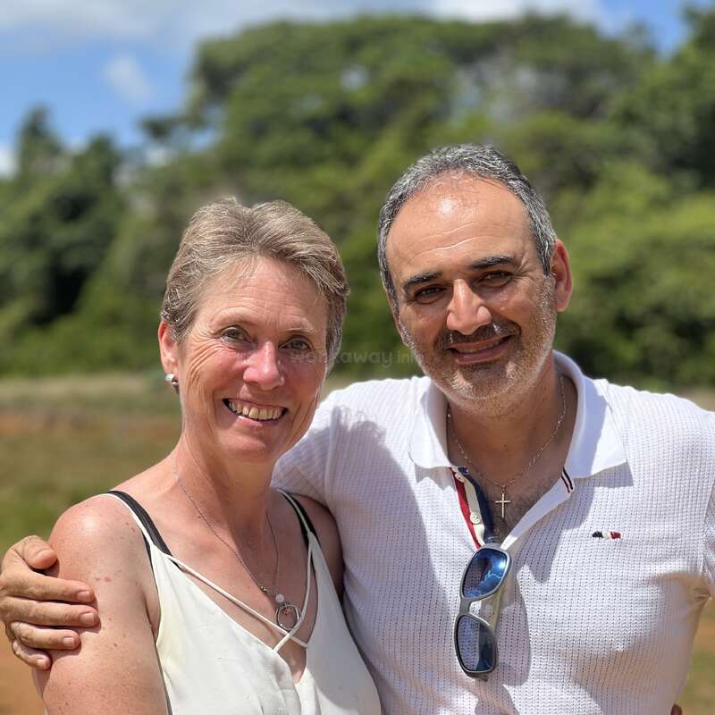 A smiling man and woman stand outdoors on a sunny day. They embrace warmly, wearing light clothing. Lush green trees and blue sky create a beautiful background.