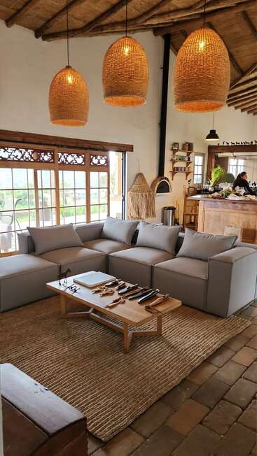 Este acogedor salón cuenta con un moderno sofá modular gris, detalles rústicos en madera, lámparas colgantes de mimbre, amplios ventanales, una alfombra de fibras naturales y una acogedora cocina abierta.