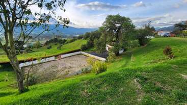 Un pintoresco paisaje rural con exuberante césped verde, árboles, un pequeño edificio blanco y una zona rectangular vallada, con montañas en la lejanía bajo un hermoso cielo azul.