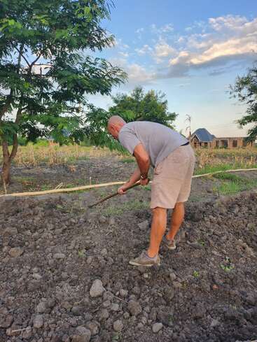 Un homme vêtu d'une chemise grise et d'un short travaille dans un jardin avec une houe, entouré d'arbres, de terre et d'un ciel partiellement nuageux.