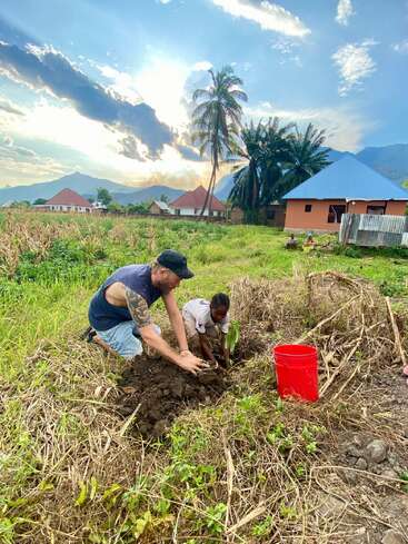 Un homme et un enfant plantent ensemble dans un champ rural au coucher du soleil, avec des maisons, des montagnes, des palmiers et un seau rouge à proximité. Le ciel est spectaculaire.