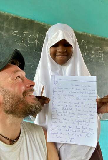 Une jeune fille souriante portant un hijab blanc montre fièrement un travail écrit à la main, debout à côté d'un homme dans une salle de classe où "STORYING" est écrit sur le tableau derrière eux.