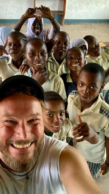 Un groupe d'écoliers souriants en uniforme pose pour un selfie joyeux avec un adulte. Tout le monde a l'air heureux, profitant du temps passé ensemble dans une salle de classe.