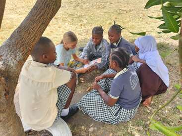 Six enfants sont assis ensemble sous un arbre, occupés à lire un livre. Ils semblent heureux et concentrés, profitant d'une expérience d'apprentissage en plein air à l'ombre.