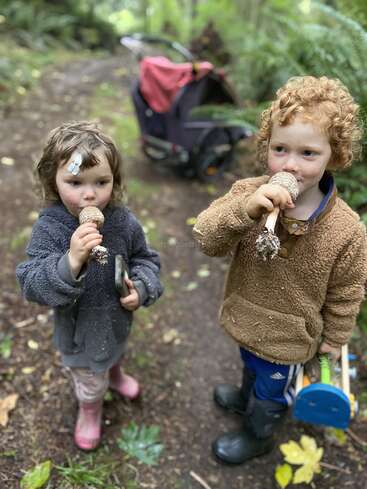 Deux jeunes enfants se tiennent sur un sentier forestier, tenant des pommes de pin contre leur bouche comme s'il s'agissait de microphones. Ils portent des vêtements chauds et des bottes, et sont entourés d'une végétation luxuriante.