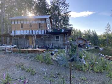 Une maison à deux étages en cours de construction se dresse au milieu des arbres, des matériaux de chantier et des fleurs sauvages. Tout près, une autre maison achevée se détache sous un ciel dégagé, évoquant un cadre champêtre.