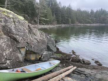 Un canoë repose sur une rive rocheuse, près de rochers recouverts de mousse. L'eau calme reflète les pins environnants, sous un ciel nuageux, créant une scène paisible et isolée.