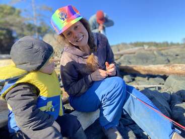 Une femme coiffée d'un chapeau coloré et vêtue d'un pantalon bleu sourit, assise sur des rochers avec un enfant bien emmitouflé dans un gilet de sauvetage. Une personne se tient debout à l'arrière-plan.