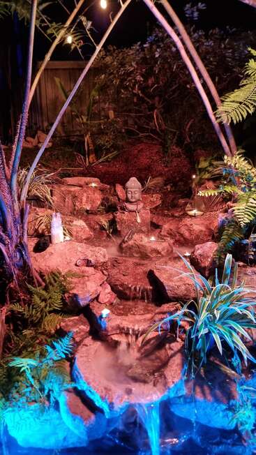 A serene garden waterfall illuminated at night, featuring a Buddha head statue, glowing candles, mist, lush green plants, and tranquil blue and purple accent lighting.