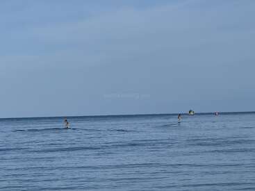 Dos personas practican paddleboarding en aguas azules y tranquilas bajo un cielo despejado. A lo lejos se ve una pequeña embarcación y una boya roja.
