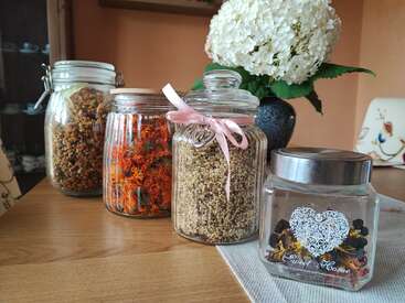 Four glass jars filled with dried herbs and flowers sit on a wooden table. In the background, there’s a beautiful white hydrangea bouquet in a vase.