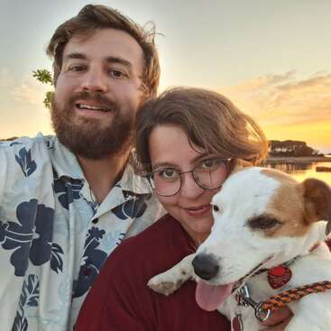 A bearded man, a woman with glasses, and a happy dog pose for a selfie outdoors near a lake at sunset, enjoying a beautiful evening.