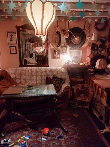 Uma sala de estar aconchegante e eclética com decoração vintage, estrelas penduradas, abajur grande, chifres, arte emoldurada, mesa desordenada, espelho, brinquedos em um carpete estampado. Iluminação quente.