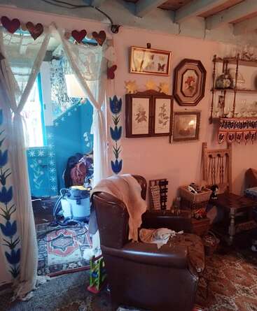 Uma sala de estar aconchegante e eclética com decoração vintage, poltrona, cortinas de renda, guirlanda de corações, pinturas florais, tapetes estampados e prateleiras cheias de pratos e bugigangas.