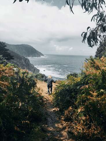Uma criança caminha por uma trilha estreita e exuberante em direção a um dramático penhasco à beira-mar. As ondas do mar batem abaixo, cercadas por vegetação e galhos pendentes.