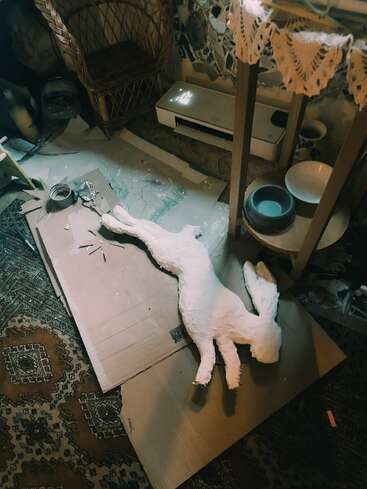 A handmade paper mache rabbit figure lies on cardboard, surrounded by art supplies, in a cozy, cluttered room with woven furniture, bowls, and patterned rug.