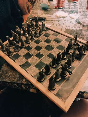 A vintage metal chess set sits on a dusty wooden board atop a patterned table. People, drinks, and scattered papers surround the cozy, game-filled atmosphere.
