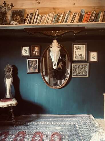 Ein gemütlicher, vielseitiger Raum mit dunklen Wänden, einem Vintage-Stuhl, einem verzierten Teppich, einem über dem Spiegel befestigten Totenkopf, gerahmten Kunstwerken und einem Regal voller Bücher.