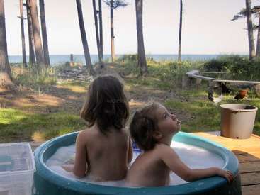 Two young children sit in a small blue pool on a wooden deck, surrounded by trees and grass, with a serene body of water in the background.