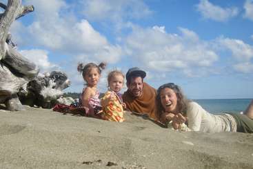 The image shows a family of four posing for a photo on the beach, with the father and mother holding their children, who are sitting on the sand. The family is dressed in casual clothing.