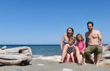 The image shows a family of four posing for a photo on the beach, with the father and mother holding their children, all dressed in matching outfits. The background is a sandy beach.