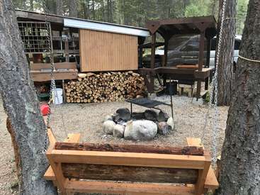 The image depicts a serene outdoor setting, featuring a fire pit surrounded by rocks, a wooden swing, and a woodpile, with a small building in the background.