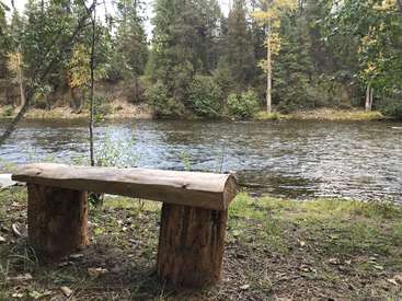 The image depicts a rustic wooden bench situated on the grassy riverbank, overlooking a serene body of water, surrounded by lush trees and foliage.
