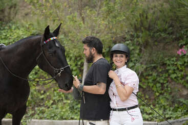 Uma mulher usando capacete está sorrindo ao lado de um cavalo preto. Um homem barbudo está ao lado do cavalo, tocando gentilmente seu rosto. Uma vegetação exuberante os rodeia.