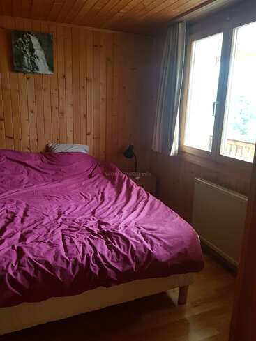 Um quarto de madeira aconchegante com uma janela grande, colcha roxa, travesseiro único, luminária de cabeceira pequena, pintura de cachoeira na parede e piso de madeira clara. A luz natural entra.