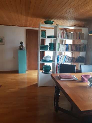 Uma sala aconchegante com teto e piso de madeira, uma estante com pratos e livros, uma mesa de jantar, cadeiras, uma estátua e luz natural suave.