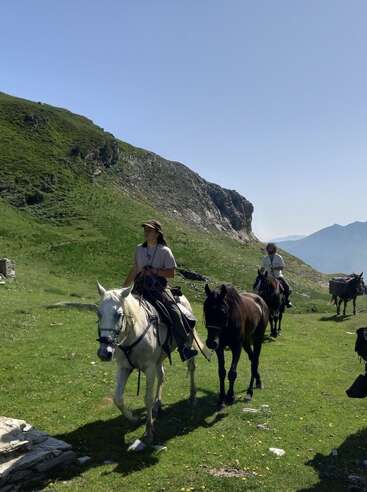 Tres personas cabalgan por un sendero montañoso cubierto de hierba bajo un cielo azul y despejado, rodeadas de verdes colinas y picos montañosos en la lejanía, disfrutando de la naturaleza.