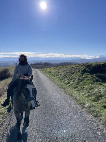 Un hombre de pelo rizado cabalga por un camino rural en un día soleado. Las majestuosas montañas y el cielo azul crean un impresionante telón de fondo.